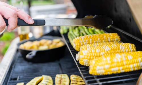 Grilling corn, zucchini, and asparagus on a BBQ grill. A hand with tongs turns the corn. A skillet with roasted potatoes is in the background.