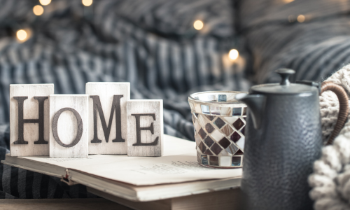 Wooden blocks spelling "HOME" on an open book, alongside a lit mosaic candle holder and rustic teapot on a cozy striped blanket, evoking warmth and comfort.