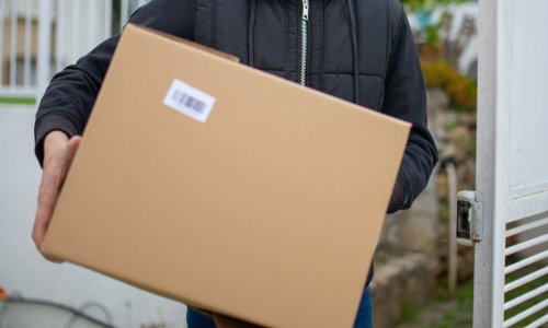 A person wearing a black jacket holds out a large cardboard package with a barcode, standing outside, suggesting a delivery or package handover.